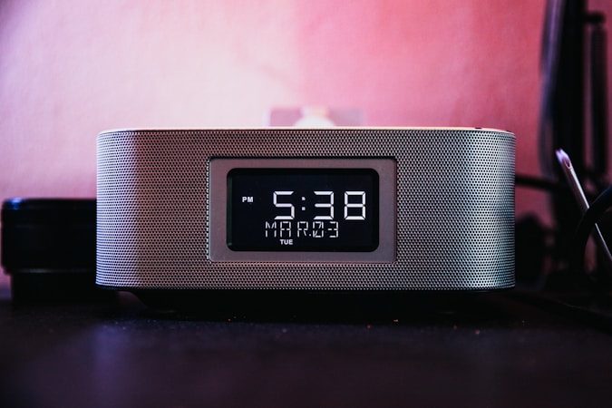 A digital clock is prominently displayed with a black and white interface, showing the time as 5:38 PM on a Tuesday, with 'Mart' mentioned below the time. The clock has a modern design with a metallic mesh exterior, placed against a pinkish background, suggesting it's on a desk or bedside table.