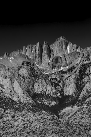 A dramatic black and white photograph of towering rugged mountain peaks, with steep, jagged rock faces and patches of snow amidst vast rocky terrain.