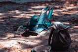 Two folding beach chairs are positioned on a sandy ground covered with dry leaves. A blue rolled-up sleeping bag rests on one of the chairs. Nearby, a gray backpack with a striped top is placed in the foreground, along with a hat on the ground. Dappled sunlight filters through, creating a pattern of light and shadow.