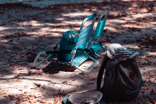 Two folding beach chairs are positioned on a sandy ground covered with dry leaves. A blue rolled-up sleeping bag rests on one of the chairs. Nearby, a gray backpack with a striped top is placed in the foreground, along with a hat on the ground. Dappled sunlight filters through, creating a pattern of light and shadow.