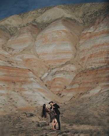 A couple is dancing in front of a vast, layered rock formation with soft shades of beige and orange creating a striking natural backdrop. The man is wearing a dark outfit and a cap, while the woman is in a floral dress. The setting appears to be a remote, arid landscape under a clear sky.