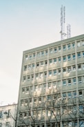 A multi-story building with numerous windows is topped with telecommunication antennas. The light gray facade is partially shadowed by nearby trees, and the clear sky forms a backdrop.