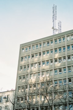 A multi-story building with numerous windows is topped with telecommunication antennas. The light gray facade is partially shadowed by nearby trees, and the clear sky forms a backdrop.