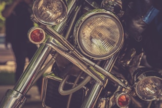 Close-up view of the front of a motorcycle, focusing on its headlight and chrome details. The image features reflections and blurred background elements, creating a vintage feel.