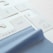 The image displays several fabric samples and garment tags on a surface. The fabric is light blue with a smooth texture, while the tags are made of white paper with printed text and icons. The text includes laundry symbols and manufacturing information.