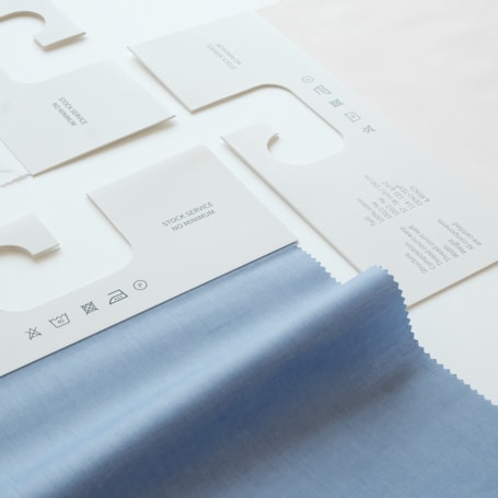The image displays several fabric samples and garment tags on a surface. The fabric is light blue with a smooth texture, while the tags are made of white paper with printed text and icons. The text includes laundry symbols and manufacturing information.