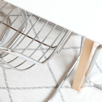 A close-up view of a metallic wire furniture piece with a wooden leg on a light-colored patterned carpet. The focus is on the sleek chrome wires contrasting with the soft texture of the fabric beneath.