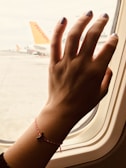 A hand with light purple nail polish stretches out towards an airplane window. The wrist has a red and white braided bracelet with a small charm. Through the window, an aircraft with a visible orange tail is parked at an airport.