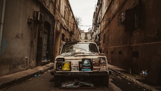 An old, deteriorating car is parked in a narrow alley between two run-down buildings. The vehicle is covered with stacks of books and various paper materials, suggesting it may be used as a street stall or storage. The setting is gritty and urban, with visible wear and decay on the surrounding architecture.