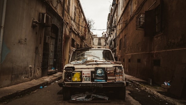 An old, deteriorating car is parked in a narrow alley between two run-down buildings. The vehicle is covered with stacks of books and various paper materials, suggesting it may be used as a street stall or storage. The setting is gritty and urban, with visible wear and decay on the surrounding architecture.