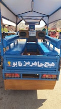 A blue open vehicle with cushioned benches on either side and a large speaker system at the front. The vehicle has a canopy overhead, with lettering in a foreign script on its back panel. There are two visible wheels and a scenic view of mountains in the background.