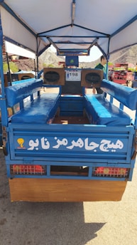 A blue open vehicle with cushioned benches on either side and a large speaker system at the front. The vehicle has a canopy overhead, with lettering in a foreign script on its back panel. There are two visible wheels and a scenic view of mountains in the background.