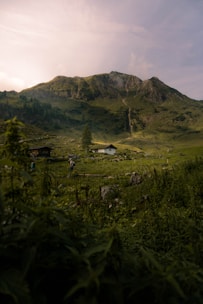 A serene mountain landscape with lush green hills and a prominent rocky peak. A white house is nestled in the valley surrounded by dense vegetation and fields. A person with a backpack walks along a path, adding a touch of activity to the tranquil scene.