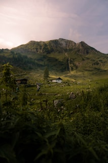 A serene mountain landscape with lush green hills and a prominent rocky peak. A white house is nestled in the valley surrounded by dense vegetation and fields. A person with a backpack walks along a path, adding a touch of activity to the tranquil scene.
