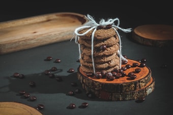 A stack of cookies tied with white string is placed on round wooden slices. Coffee beans are scattered around, adding a rustic touch. The lighting creates soft shadows, emphasizing the textures of the cookies and wood.