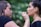 A woman applies makeup to another woman in an outdoor setting with a blurred, green leafy background. The makeup artist is focused on applying lip makeup with a small brush, while the person receiving makeup has an expression of calm concentration.
