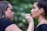 A woman applies makeup to another woman in an outdoor setting with a blurred, green leafy background. The makeup artist is focused on applying lip makeup with a small brush, while the person receiving makeup has an expression of calm concentration.