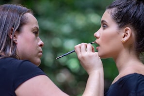 A woman applies makeup to another woman in an outdoor setting with a blurred, green leafy background. The makeup artist is focused on applying lip makeup with a small brush, while the person receiving makeup has an expression of calm concentration.