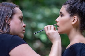 A woman applies makeup to another woman in an outdoor setting with a blurred, green leafy background. The makeup artist is focused on applying lip makeup with a small brush, while the person receiving makeup has an expression of calm concentration.