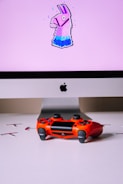 A computer monitor displays an image of a stylized llama with a pink background. In front of the monitor, a vibrant red gaming controller sits on a white surface.