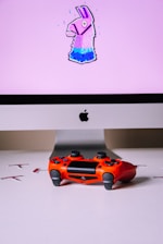 A computer monitor displays an image of a stylized llama with a pink background. In front of the monitor, a vibrant red gaming controller sits on a white surface.