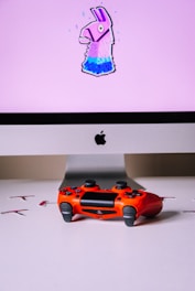 A computer monitor displays an image of a stylized llama with a pink background. In front of the monitor, a vibrant red gaming controller sits on a white surface.