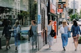 Three women are walking along a city street, each carrying a backpack. They are passing by a glass storefront that reflects their image. The sidewalk is lined with buildings displaying signs in Cyrillic script, and some advertisements offer exchange rates for currencies. The scene is urban and features typical city elements like shops and street signs.