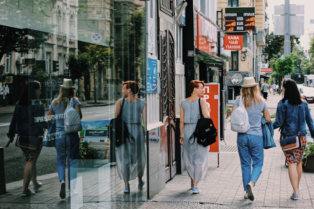 Three women are walking along a city street, each carrying a backpack. They are passing by a glass storefront that reflects their image. The sidewalk is lined with buildings displaying signs in Cyrillic script, and some advertisements offer exchange rates for currencies. The scene is urban and features typical city elements like shops and street signs.