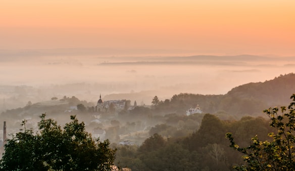 A serene landscape with a village nestled among rolling hills, shrouded in early morning mist. The sky transitions from a soft orange hue at the horizon to a pale blue as it rises. Trees and foliage frame the foreground, while a church with a spire stands out in the distant village.