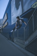 A person is performing a parkour move, jumping over a metal railing beside a large, blue building with IKEA signage. The person is dressed casually in a t-shirt and jeans with sneakers, displaying athleticism. There is a billboard advertisement visible on the wall of the building.