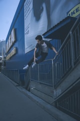 A person is performing a parkour move, jumping over a metal railing beside a large, blue building with IKEA signage. The person is dressed casually in a t-shirt and jeans with sneakers, displaying athleticism. There is a billboard advertisement visible on the wall of the building.