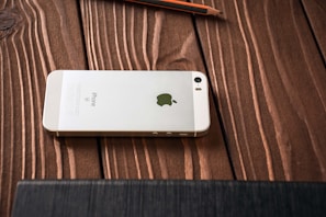 A white smartphone with an Apple logo on its back is placed face down on a wooden surface. The wooden grain texture is visible, and a pencil is lying nearby.