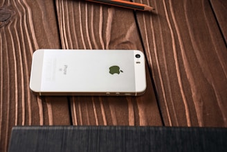 A white smartphone with an Apple logo on its back is placed face down on a wooden surface. The wooden grain texture is visible, and a pencil is lying nearby.
