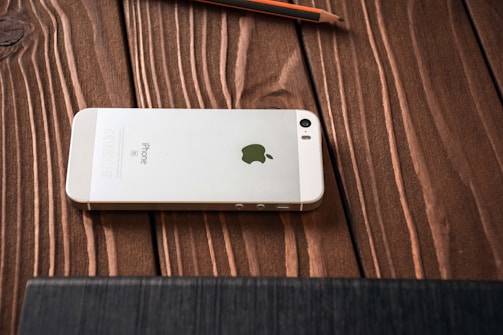 A white smartphone with an Apple logo on its back is placed face down on a wooden surface. The wooden grain texture is visible, and a pencil is lying nearby.