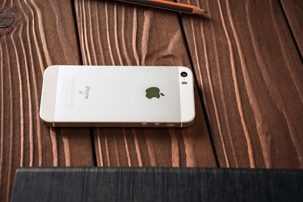 A white smartphone with an Apple logo on its back is placed face down on a wooden surface. The wooden grain texture is visible, and a pencil is lying nearby.