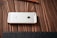 A white smartphone with an Apple logo on its back is placed face down on a wooden surface. The wooden grain texture is visible, and a pencil is lying nearby.