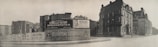 A black-and-white photograph of an urban street corner featuring a large advertising billboard. The billboard displays ads for Stewart Speedometer, Beech-Nut Sliced Bacon, and Hunter Whiskey. Surrounding the billboard are older, multi-story buildings with classic architecture.