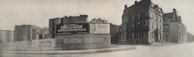 A black-and-white photograph of an urban street corner featuring a large advertising billboard. The billboard displays ads for Stewart Speedometer, Beech-Nut Sliced Bacon, and Hunter Whiskey. Surrounding the billboard are older, multi-story buildings with classic architecture.