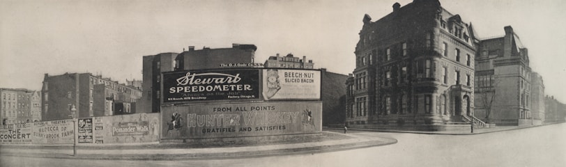 A black-and-white photograph of an urban street corner featuring a large advertising billboard. The billboard displays ads for Stewart Speedometer, Beech-Nut Sliced Bacon, and Hunter Whiskey. Surrounding the billboard are older, multi-story buildings with classic architecture.