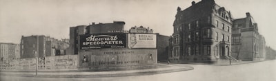 A black-and-white photograph of an urban street corner featuring a large advertising billboard. The billboard displays ads for Stewart Speedometer, Beech-Nut Sliced Bacon, and Hunter Whiskey. Surrounding the billboard are older, multi-story buildings with classic architecture.