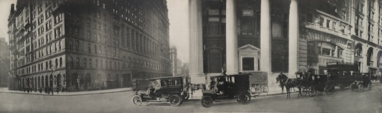 A vintage street scene features early 20th-century architecture with tall buildings in a city setting. Horse-drawn carriages and early automobiles are parked on the street, indicating a bustling urban environment.