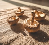 Four brown mushrooms are placed on a textured, woven surface with sunlight casting soft shadows. The mushrooms have visible stems and smooth caps, and they appear to be fresh.