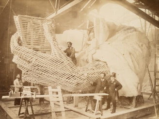 A group of men is working on the construction of a large-scale sculpture, focusing on a section resembling a hand. The framework and detailing of the sculpture are visible, with some workers positioned on ladders and scaffolding. The environment appears to be an indoor workshop with high ceilings.