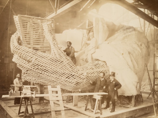 A group of men is working on the construction of a large-scale sculpture, focusing on a section resembling a hand. The framework and detailing of the sculpture are visible, with some workers positioned on ladders and scaffolding. The environment appears to be an indoor workshop with high ceilings.
