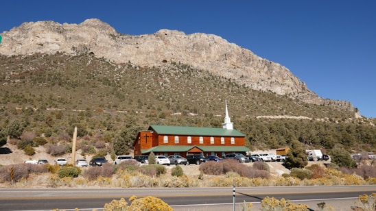 A church with a white steeple and green roof stands at the foot of a large, rocky mountain. Several cars are parked in front of the church, surrounded by sparse vegetation and desert shrubs. The sky is clear and blue, creating a stark contrast with the rugged terrain of the mountain.