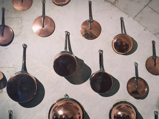 A collection of copper pans of various sizes with long handles hanging on a textured white wall. The pans have a shiny, reflective surface, casting distinct shadows, and some have a polished appearance. The arrangement creates a visually appealing pattern.