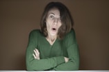 A person with short brown hair, wearing a green sweater, has a surprised expression on their face with raised eyebrows and open mouth. The arms are crossed on a white surface, and the background is plain brown.