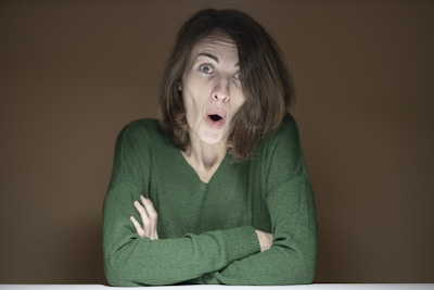 A person with short brown hair, wearing a green sweater, has a surprised expression on their face with raised eyebrows and open mouth. The arms are crossed on a white surface, and the background is plain brown.