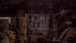 A dimly lit art gallery displays various sculptures and artifacts in glass cases. A person is observing one of the exhibits, surrounded by intricately carved wooden pieces with patterns and figures. The atmosphere is quiet and contemplative.