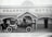 A vintage car with passengers is parked in front of an old-fashioned hotel named Shamrock Hotel. The hotel has a facade with striped awnings and ornate woodwork. Four men are in the car, and two men are standing nearby, dressed in early 20th-century attire. The name P. Sweeney is visible on the building.